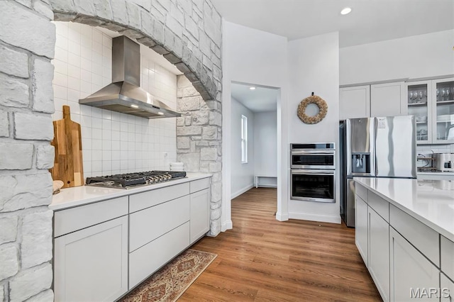 213 Bluegrass Pasture Ct, Wentzville, MO 63385 - kitchen with wall chimney exhaust hood, appliances with stainless steel finishes, backsplash, glass insert cabinets, and wood finished floors