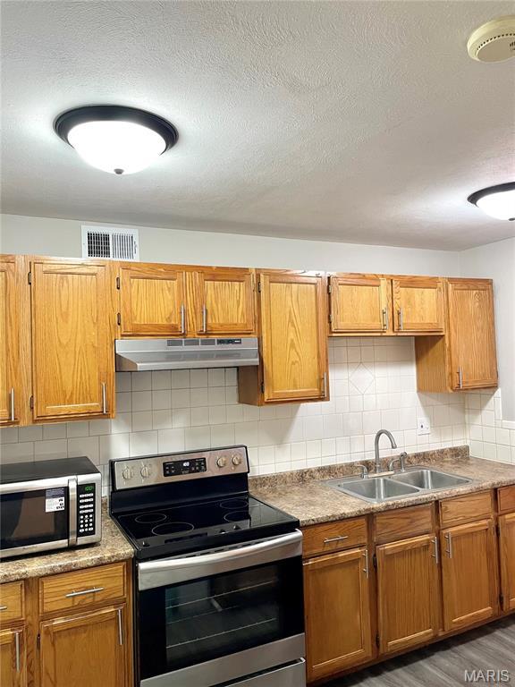 9264 Bellefontaine Rd, Bellefontaine Nghbrs, MO 63137 - kitchen featuring appliances with stainless steel finishes, under cabinet range hood, backsplash, a textured ceiling, and brown cabinets