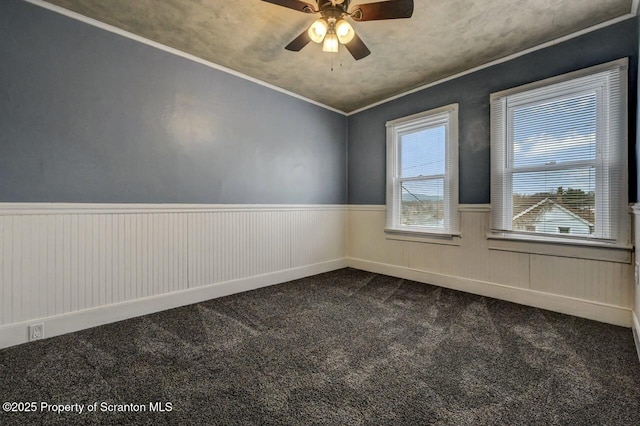 empty room featuring wainscoting, crown molding, ceiling fan, and dark colored carpet