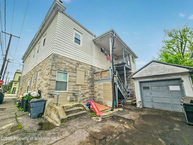 view of side of home featuring stone siding, a garage, aphalt driveway, a balcony, and an outbuilding