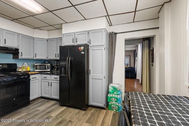 kitchen with black appliances, under cabinet range hood, light wood finished floors, and a drop ceiling