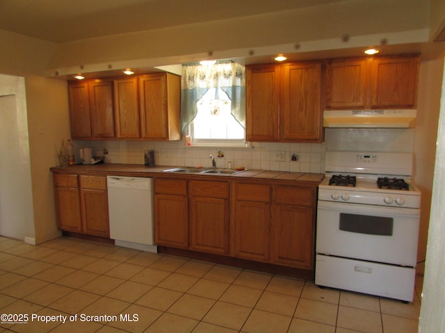 kitchen with tile counters, under cabinet range hood, brown cabinets, white appliances, and a sink