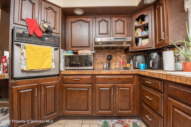 kitchen featuring under cabinet range hood, stainless steel appliances, tile countertops, light tile patterned flooring, and decorative backsplash