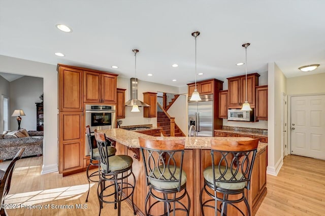 kitchen featuring wall chimney range hood, an island with sink, appliances with stainless steel finishes, decorative light fixtures, and light stone counters