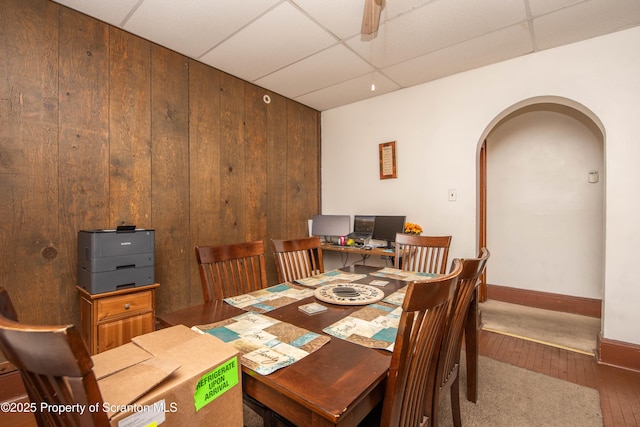 dining area featuring wooden walls, baseboards, arched walkways, a paneled ceiling, and wood-type flooring