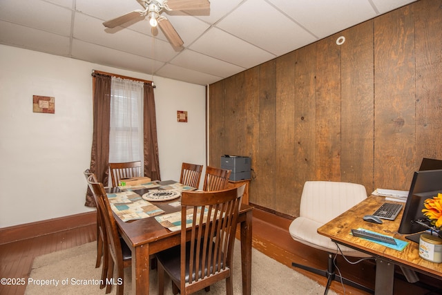 dining area featuring a drop ceiling, ceiling fan, wooden walls, and wood finished floors