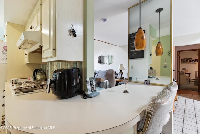 kitchen with light countertops, tile patterned flooring, under cabinet range hood, cream cabinetry, and hanging light fixtures