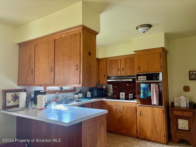 kitchen featuring brown cabinetry, a peninsula, under cabinet range hood, light countertops, and black appliances