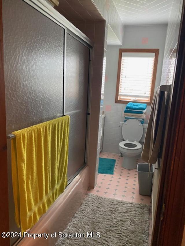 bathroom with toilet, enclosed tub / shower combo, and tile walls