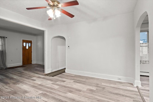 unfurnished living room featuring ceiling fan, arched walkways, baseboard heating, baseboards, and light wood-style floors