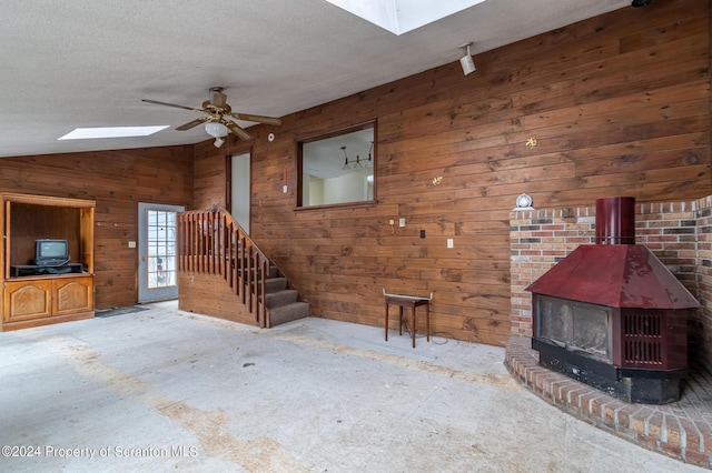 unfurnished living room with a textured ceiling, lofted ceiling with skylight, ceiling fan, a wood stove, and wood walls