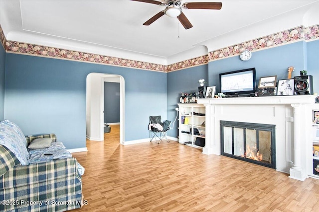 living area with arched walkways, a ceiling fan, a glass covered fireplace, wood finished floors, and baseboards