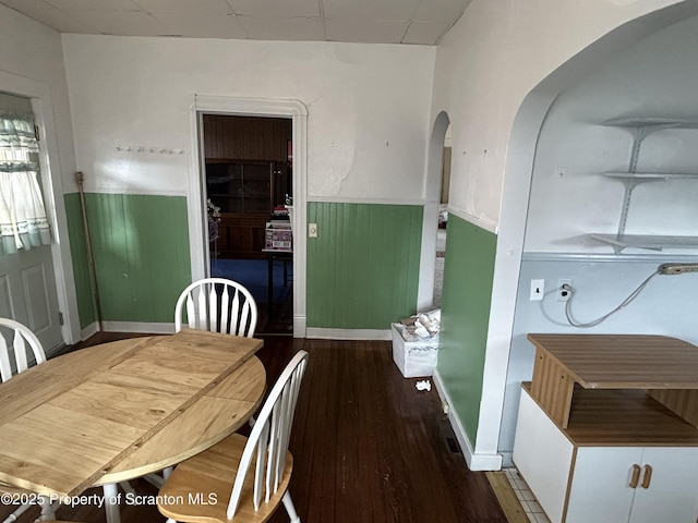 dining space featuring dark wood-type flooring, arched walkways, wainscoting, and a drop ceiling