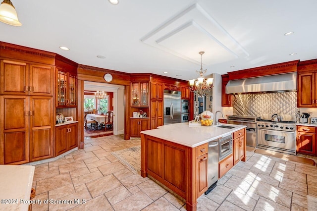 kitchen with wall chimney exhaust hood, a kitchen island with sink, built in appliances, a chandelier, and hanging light fixtures