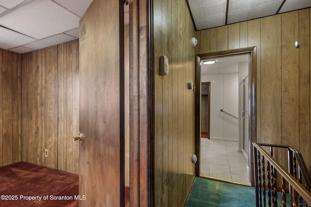 corridor featuring wooden walls, carpet floors, tile patterned flooring, a paneled ceiling, and an upstairs landing