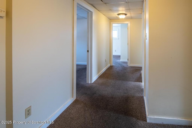 hallway with baseboards, a paneled ceiling, and dark carpet