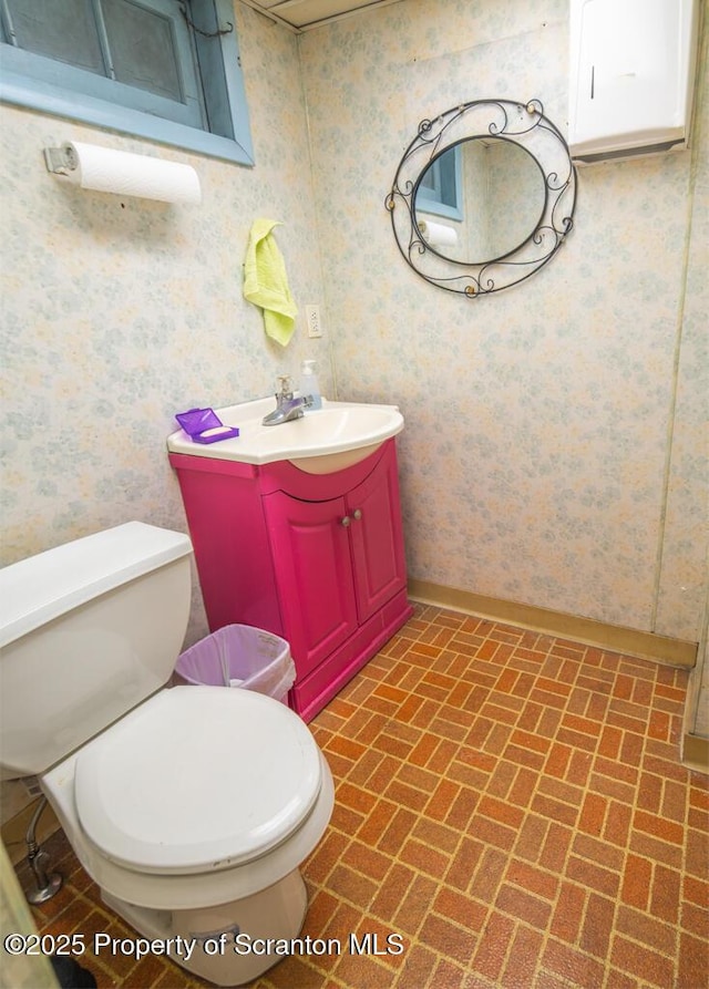 half bathroom with vanity, baseboards, wallpapered walls, brick floor, and toilet