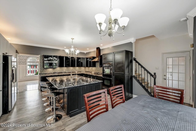 kitchen with a center island with sink, wall chimney range hood, decorative backsplash, appliances with stainless steel finishes, and a notable chandelier