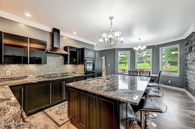 kitchen with stone counters, wall chimney range hood, an inviting chandelier, decorative light fixtures, and a kitchen island with sink