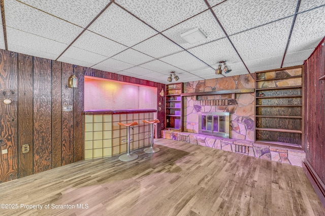 unfurnished living room featuring a fireplace, wood walls, wood-type flooring, a drop ceiling, and built in shelves