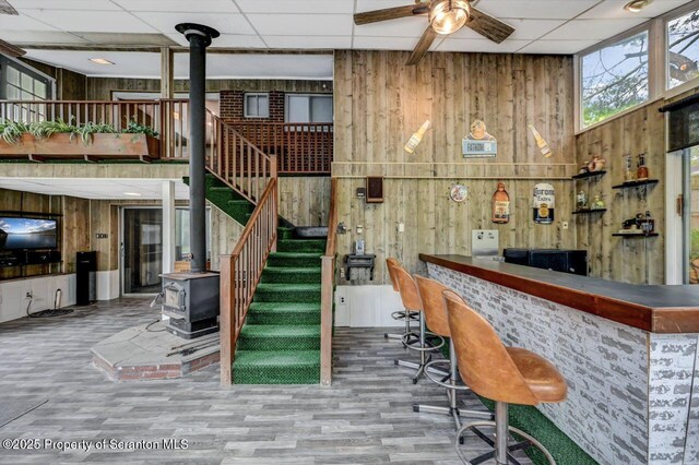 indoor dry bar featuring a wood stove, a paneled ceiling, wood walls, wood finished floors, and ceiling fan