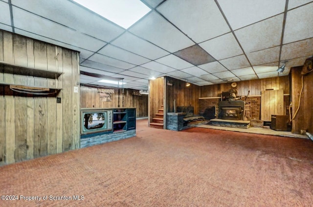 unfurnished living room with a paneled ceiling, wood walls, a wood stove, and carpet