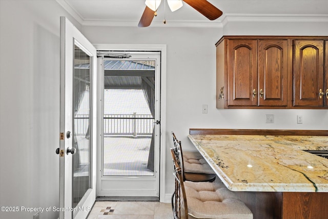 kitchen with light tile patterned floors, brown cabinetry, ceiling fan, ornamental molding, and light stone countertops