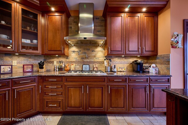 kitchen with wall chimney exhaust hood, dark stone countertops, decorative backsplash, and stainless steel gas cooktop