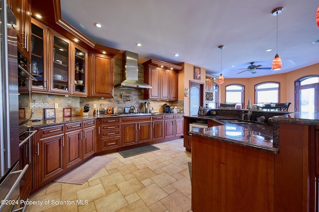 kitchen with hanging light fixtures, wall chimney exhaust hood, ceiling fan, dark stone countertops, and tasteful backsplash