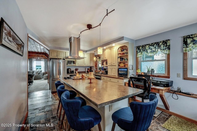 kitchen with appliances with stainless steel finishes, a center island, island range hood, cream cabinetry, and hanging light fixtures