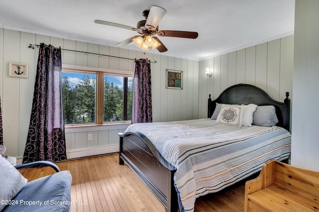 bedroom with ceiling fan, light wood-type flooring, baseboard heating, and wooden walls