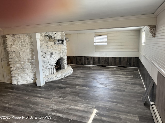 unfurnished living room with ornamental molding, a baseboard heating unit, wood finished floors, wooden walls, and a stone fireplace