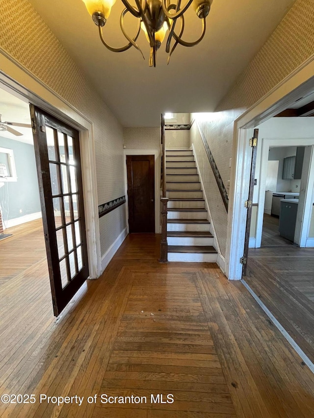 hall with dark wood-style floors, stairway, a chandelier, baseboards, and wallpapered walls