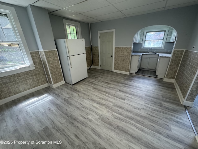 unfurnished dining area with wainscoting, a paneled ceiling, tile walls, light wood finished floors, and arched walkways