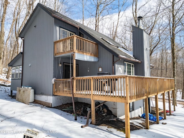 snow covered back of property featuring a deck, roof with shingles, a chimney, and a balcony