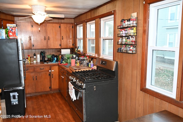 kitchen featuring stainless steel gas stove, wooden walls, brown cabinetry, dark wood finished floors, and freestanding refrigerator