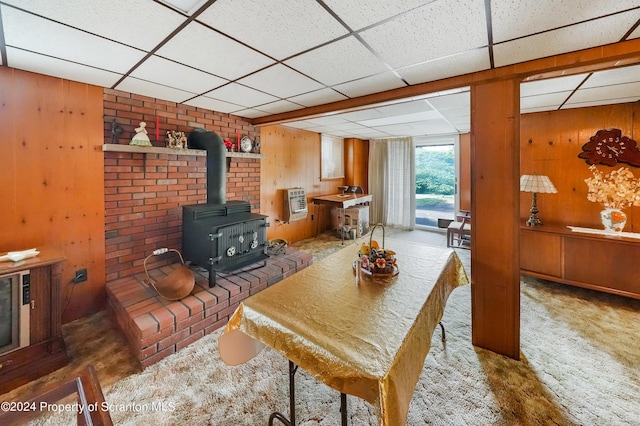 carpeted living room featuring a drop ceiling, a wood stove, wooden walls, and heating unit