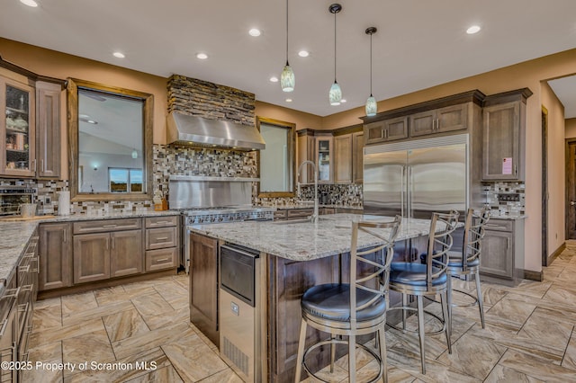 815 Oak St, Archbald, PA 18403 - kitchen with an island with sink, light stone counters, built in fridge, wall chimney range hood, and glass insert cabinets