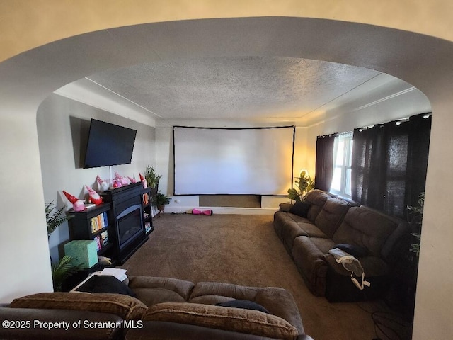 carpeted cinema room featuring arched walkways, a glass covered fireplace, and a textured ceiling