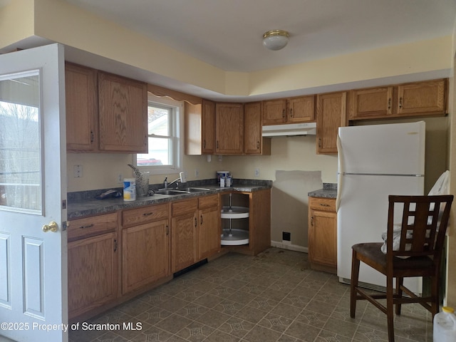 kitchen featuring a sink, under cabinet range hood, dark countertops, freestanding refrigerator, and brown cabinetry