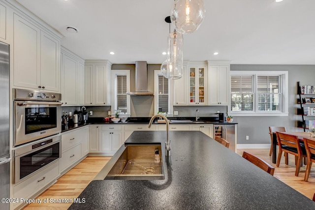 kitchen featuring wall chimney exhaust hood, stainless steel appliances, light hardwood / wood-style flooring, white cabinets, and hanging light fixtures