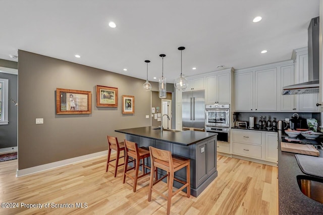 kitchen with wall chimney exhaust hood, a breakfast bar area, a center island with sink, appliances with stainless steel finishes, and light wood-type flooring