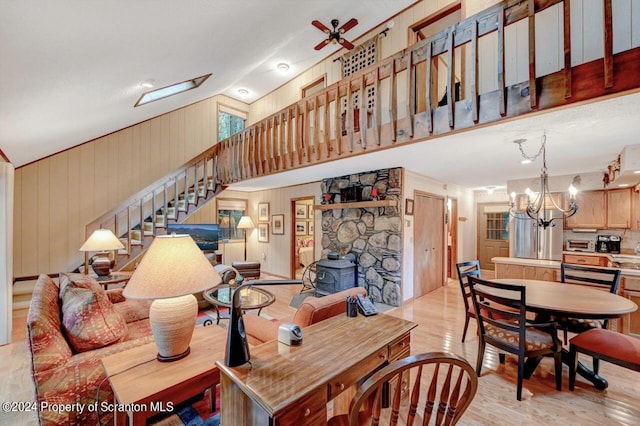 living room with ceiling fan with notable chandelier, a wood stove, wooden walls, and vaulted ceiling