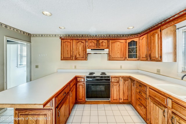 kitchen with a sink, a peninsula, under cabinet range hood, brown cabinetry, and black electric range