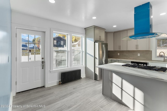 kitchen featuring appliances with stainless steel finishes, plenty of natural light, island range hood, light stone counters, and radiator heating unit