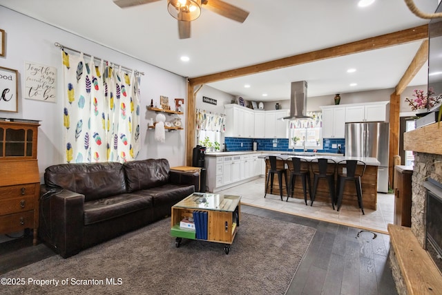 living area with a ceiling fan, a stone fireplace, beamed ceiling, light wood finished floors, and recessed lighting
