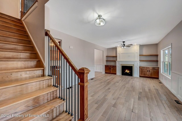 unfurnished living room featuring a warm lit fireplace, ceiling fan, stairway, and light wood-style flooring