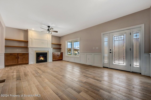 unfurnished living room with a decorative wall, a ceiling fan, wainscoting, light wood-type flooring, and a glass covered fireplace