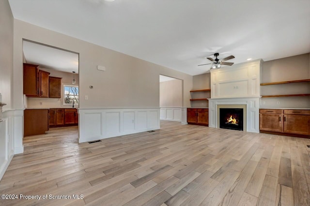 unfurnished living room with a ceiling fan, wainscoting, a lit fireplace, light wood-style floors, and a decorative wall