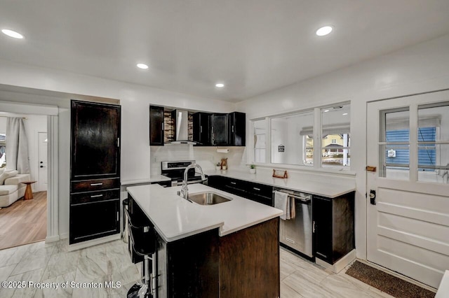 kitchen with stainless steel appliances, a sink, light countertops, wall chimney range hood, and an island with sink